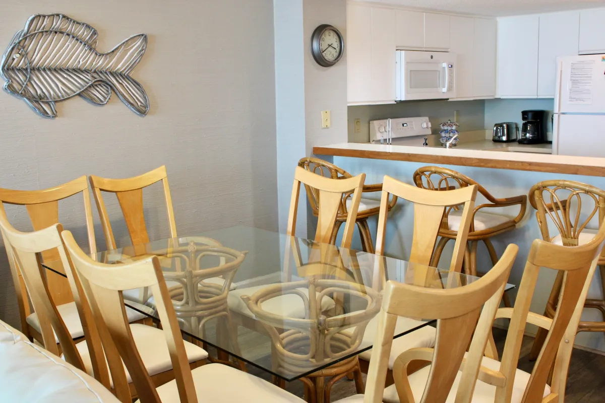 A light and airy interior with a glass dining table surrounded by wooden chairs, fish wall art, and a visible kitchen area in the background. The space feels designed for family gatherings and shared meals.
