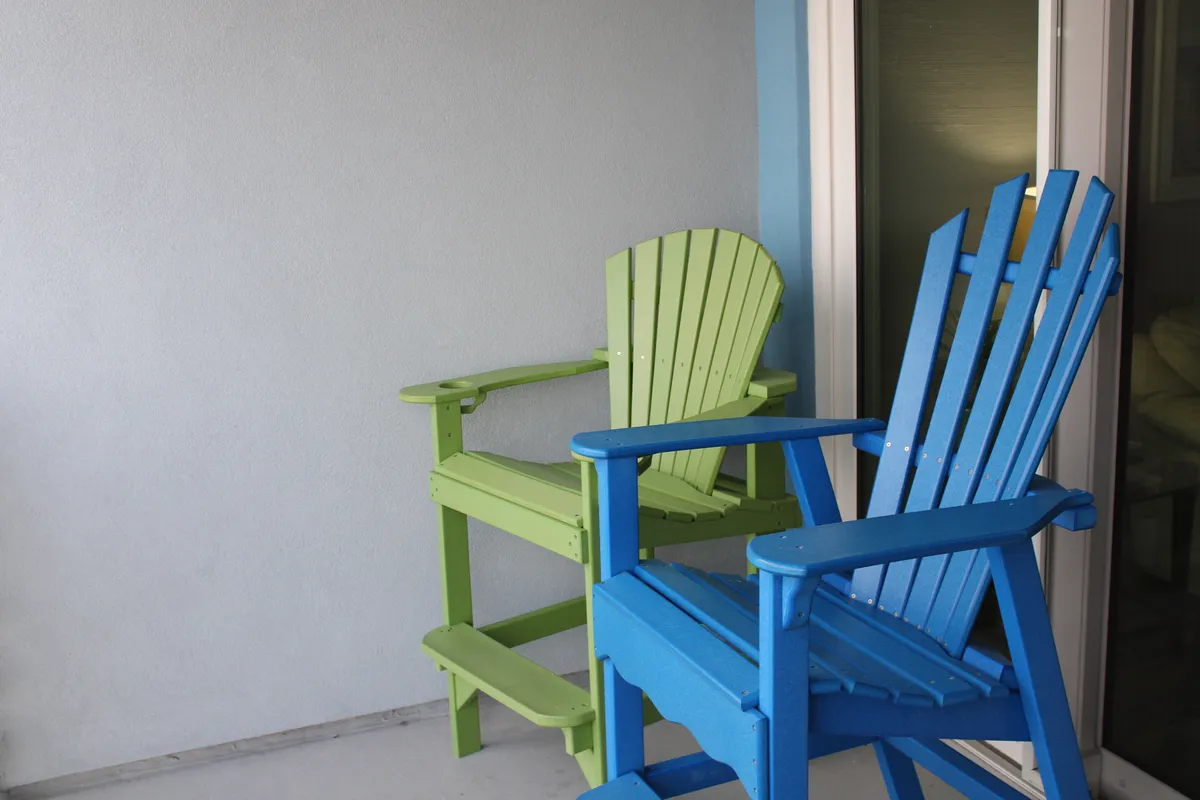 A close-up view of green and blue Adirondack chairs on a balcony, evoking a sense of comfort and tranquility. The composition focuses on the inviting seating setup, ideal for quiet moments or lively gatherings with the ocean atmosphere just beyond.