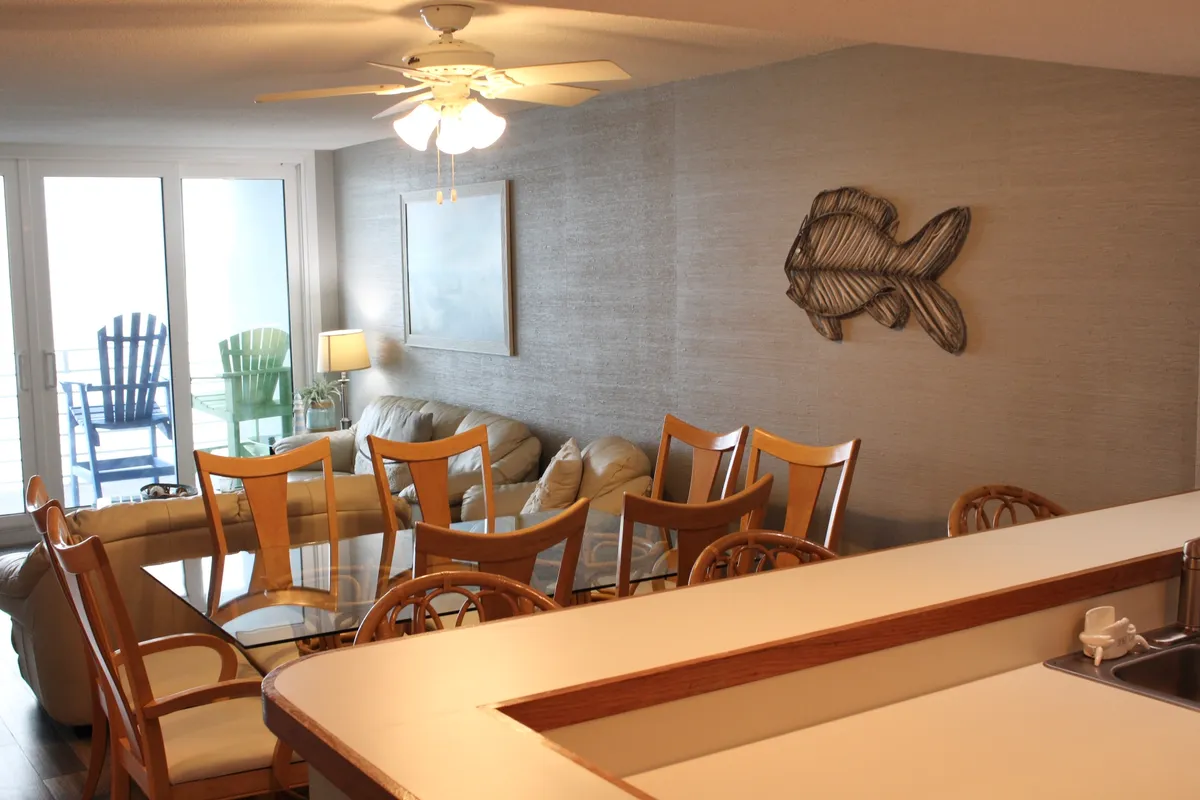 This image showcases a comfortable open-plan area featuring a dining table with chairs, a sofa, decorative fish wall art, and large windows that offer views of outdoor seating. The natural light floods the space, creating an inviting atmosphere for gatherings.