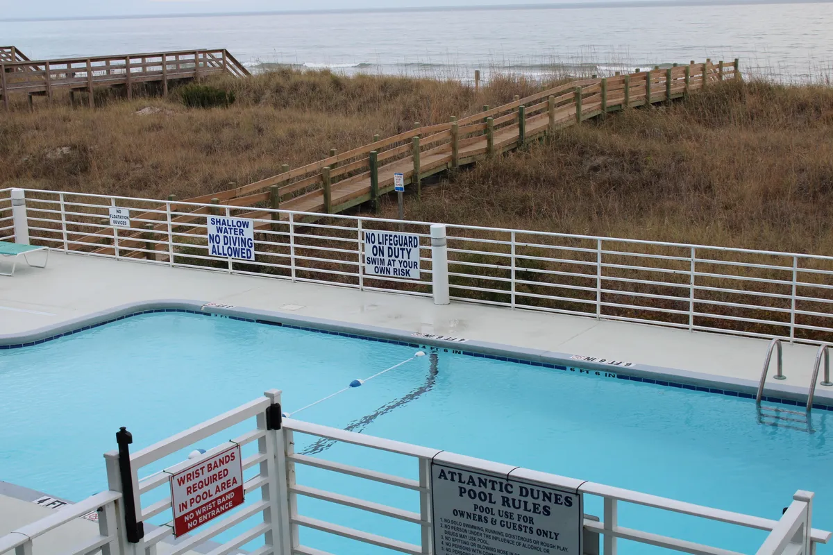 A calm pool area with a swimming pool in the foreground, a wooden walkway leading toward a beachfront view, pool rules signs, and the beach and ocean visible in the distance. The tranquil ambiance and broad perspective capture the seamless transition from pool to beach.