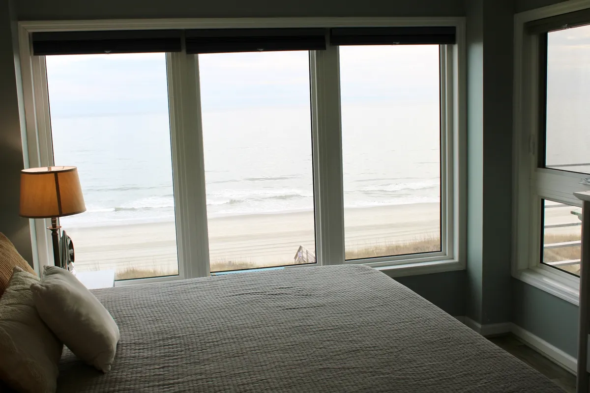 This image features a tranquil bedroom with large windows that frame sweeping views of the beach. The comfortable bed is topped with plush pillows, while a table lamp creates a cozy ambiance. The scene captures the serene experience of waking up to the ocean just steps away.