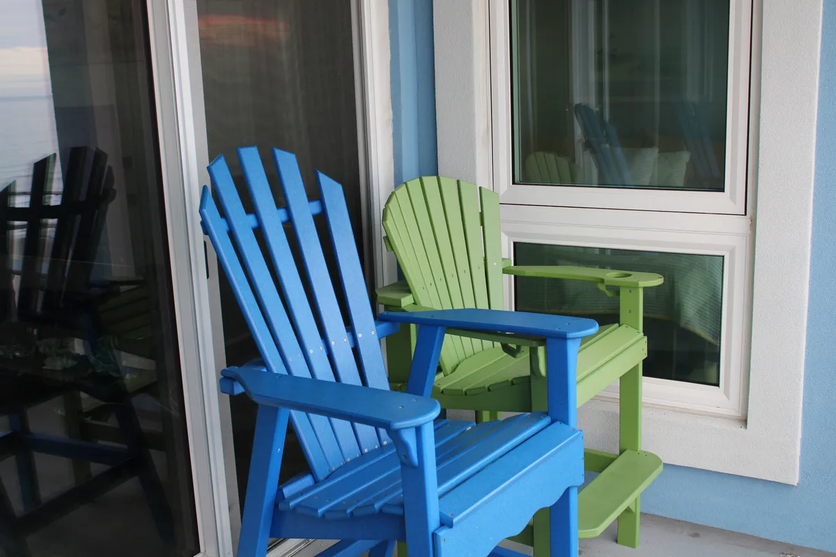 A pair of blue and green Adirondack chairs positioned on a balcony, oriented to capture a relaxing view beyond the balcony railing. The window behind hints at a seamless indoor-outdoor transition and suggests a welcoming space for enjoying the scenery.