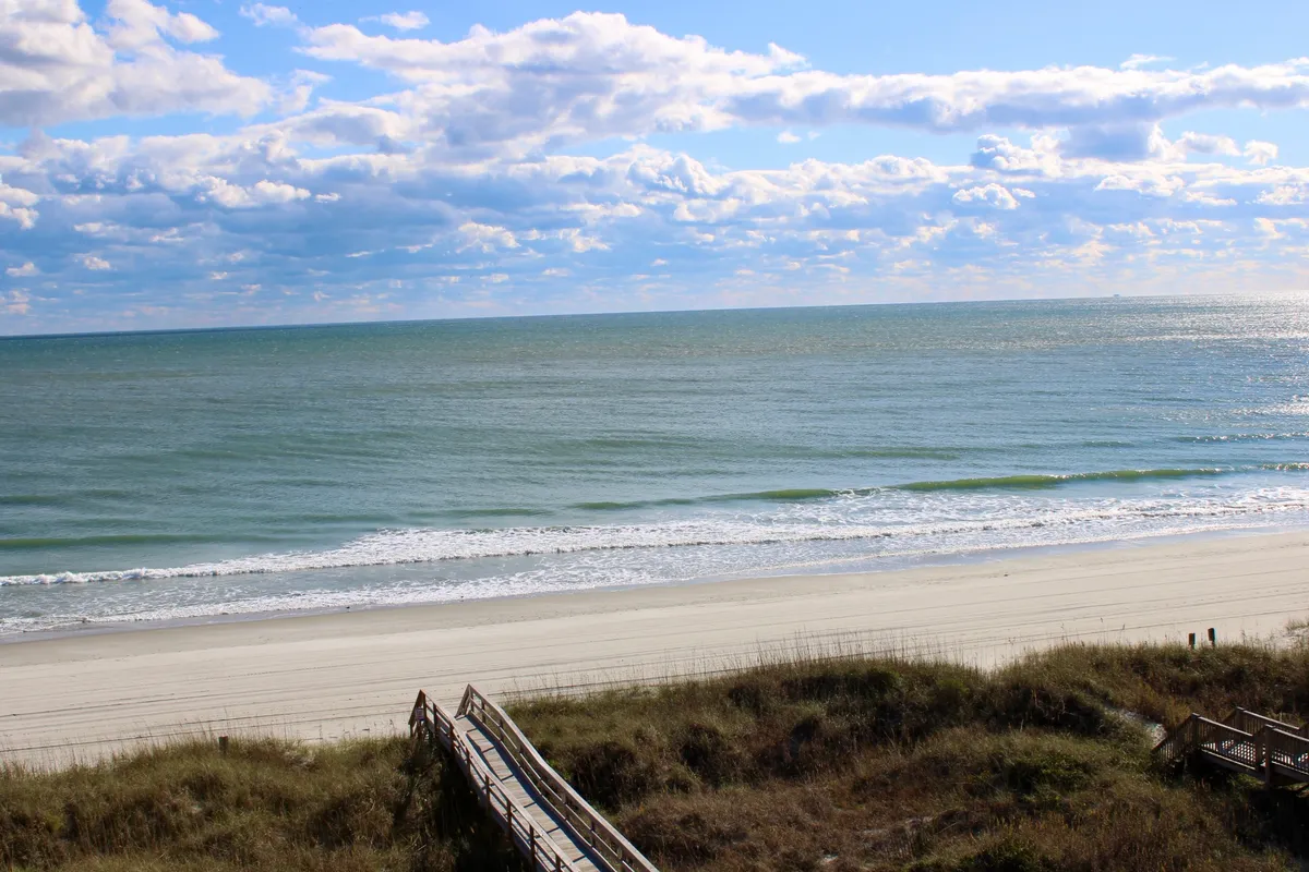 The image showcases a breathtaking oceanfront view from the condo, with a private boardwalk leading to the sandy beach. The gentle waves of the Atlantic stretch out to the horizon beneath a sky brushed with soft clouds. The scene evokes tranquility, with inviting space for lounging on the sand, walking along the shore, and watching unforgettable sunsets.