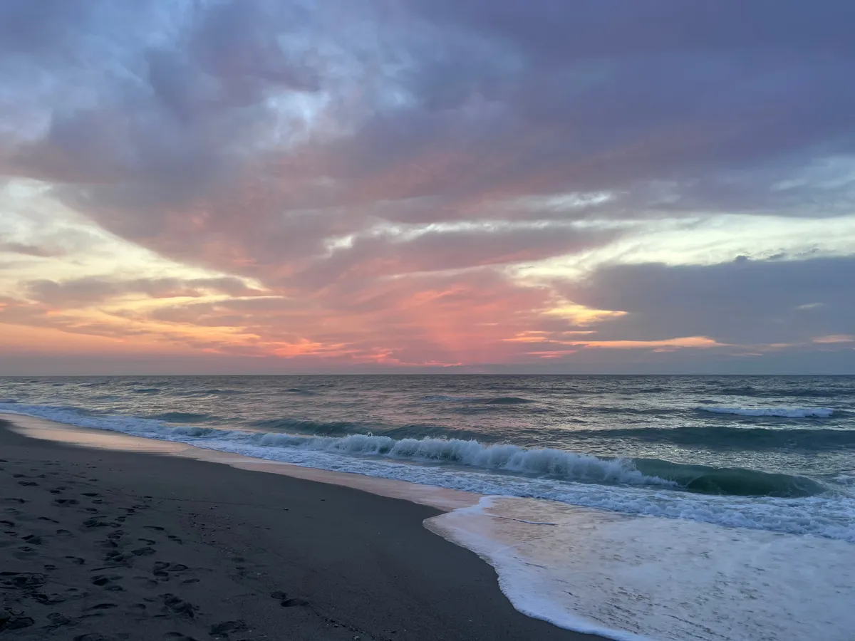 A tranquil exterior shot showing a sandy beach at sunset, with gentle waves rolling in and a picturesque sky painted in sunset hues. The scene evokes peaceful evening walks and memorable moments by the water.