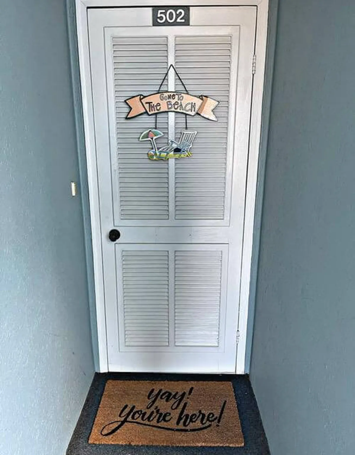 A cheerful entryway with a beach-themed sign, a welcoming mat, and the condo’s door number, evoking the excitement of arriving at a vacation home right by the ocean. The decor hints at the coastal lifestyle awaiting guests inside.