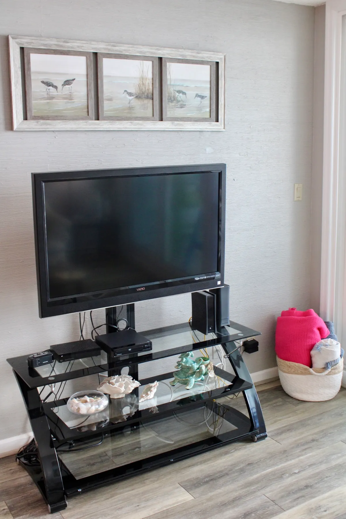 A cozy living area featuring a television, framed artwork, decorative shelving, and a basket with blankets. The setting is arranged for comfort and entertainment, suggesting a space for movie nights or board games.