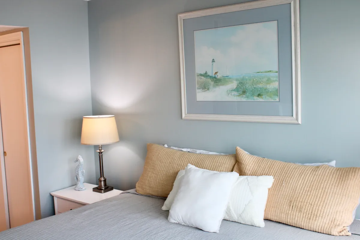 A serene bedroom scene showcasing a neatly made bed with decorative pillows, a bedside lamp, and framed artwork. The soft, inviting palette and gentle lighting create a cozy, restful atmosphere, perfect for unwinding.