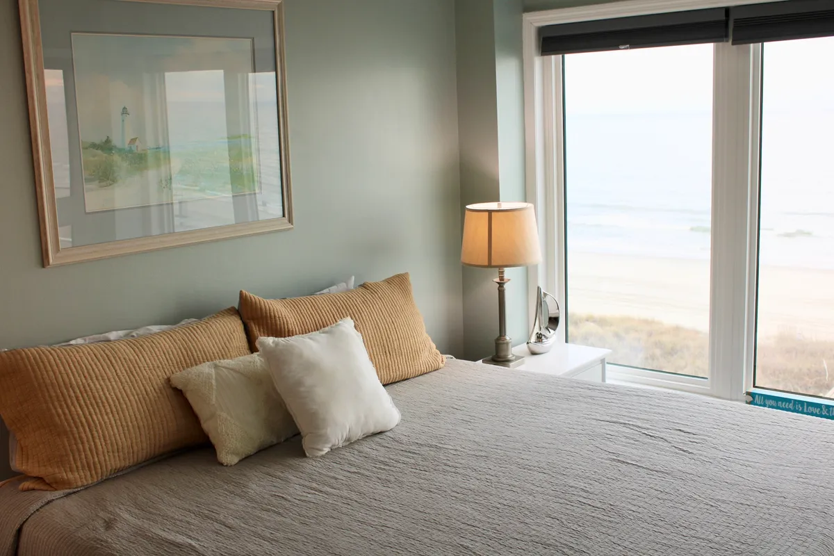 A tranquil bedroom space featuring a plush bed with textured pillows, a large window framing an ocean view, a bedside lamp, and calm, coastal-inspired framed artwork. Soft natural light fills the room, highlighting the cozy bedding and peaceful aesthetic.