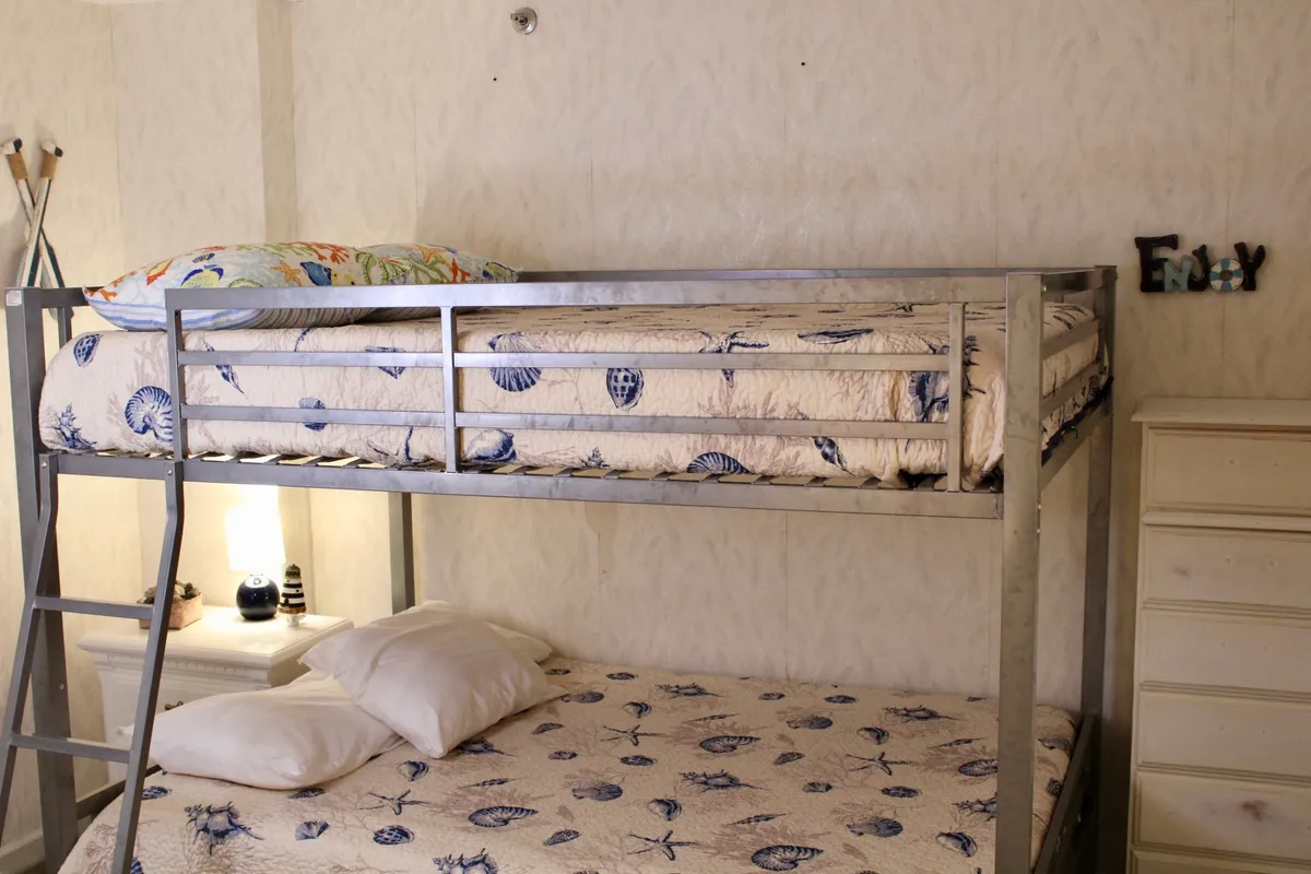A cheerful bedroom furnished with two sets of full-size bunk beds, decorative pillows, a nightstand with a lamp, and a chest of drawers. The playful yet inviting design welcomes kids, teens, or friends to relax, chat, and enjoy sleepovers.