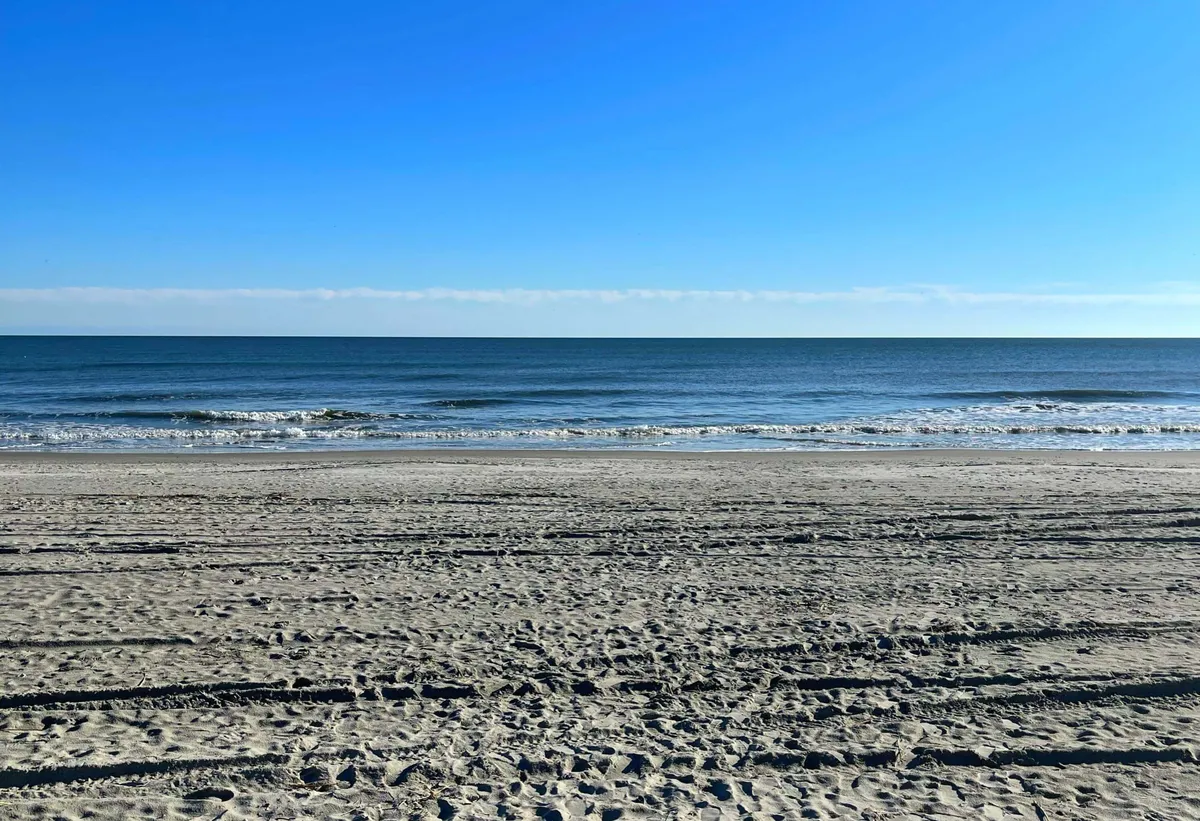 A wide stretch of sandy beach meets gentle ocean waves under a clear sky, offering ample space for sunbathing, swimming, and beachcombing. The serene setting invites guests to build sandcastles, enjoy the water, or simply soak up the sun.