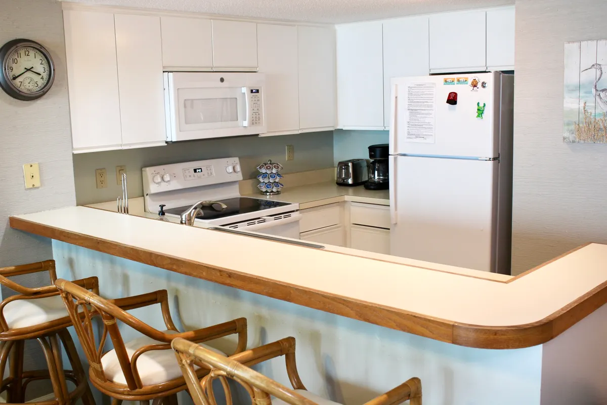 A bright kitchen scene with a counter and bar stools, white cabinetry, and visible kitchen appliances. The layout encourages socializing and cooking together, highlighting the functional yet welcoming nature of the kitchen.