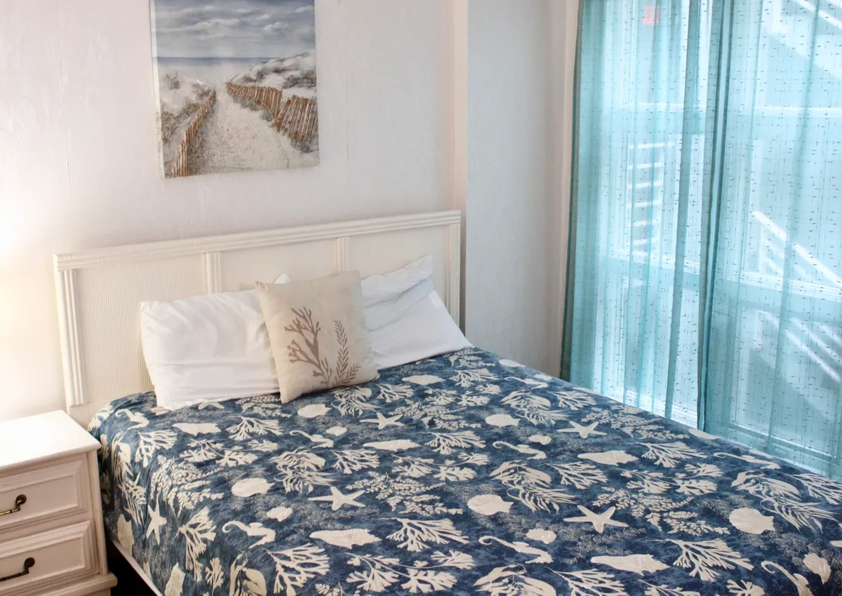 A serene bedroom scene with a coastal-themed blanket, decorative marine pillows, and artwork depicting a beach scene. Sheer turquoise curtains allow natural light to gently filter in, enhancing the calming atmosphere. The window offers glimpses of the changing light at sunrise or sunset, echoing the promise of romantic and tranquil moments.